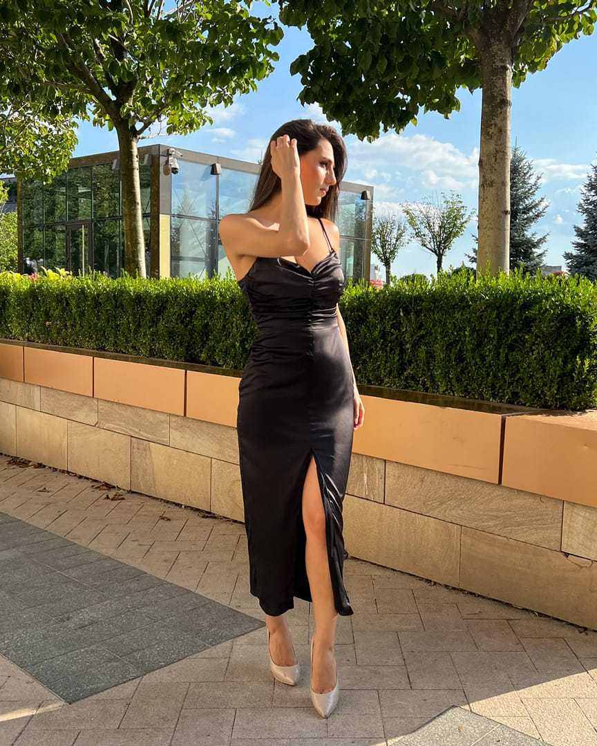 Rochie cocktail neagră de seară din satin elastic cu tăietură centrală și pliuri, purtată de un model.